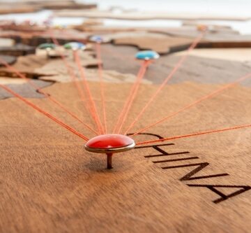 A wooden world map with a red flag pin stuck into the area labeled "CHINA" with red threads extending from the central pin in China to various other points on the map. A wooden world map with a red flag pin stuck into the area labeled "CHINA" with red threads extending from the central pin in China to various other points on the map.
