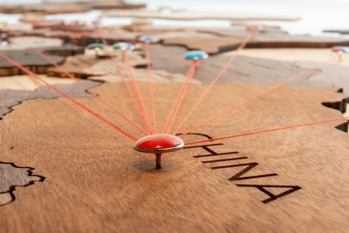 A wooden world map with a red flag pin stuck into the area labeled "CHINA" with red threads extending from the central pin in China to various other points on the map. A wooden world map with a red flag pin stuck into the area labeled "CHINA" with red threads extending from the central pin in China to various other points on the map.