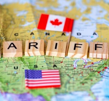 A row of wooden tiles spelling "TARIFFS" laid across a map of Canada and the U.S. with miniature flags atop each country. A row of wooden tiles spelling "TARIFFS" laid across a map of Canada and the U.S. with miniature flags atop each country.