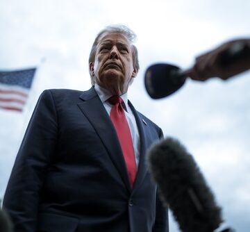Donald Trump addressing the press in a low-angle shot with microphones in the foreground and an American flag visible in the background. Donald Trump addressing the press in a low-angle shot with microphones in the foreground and an American flag visible in the background.