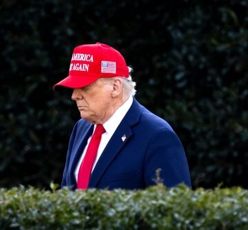 Donald Trump, wearing a red "Make America Great Again" hat and a blue suit with a red tie, while walking near a hedge on the White House South Lawn.