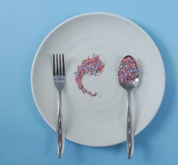 A white plate against a blue background with a fork and a spoon holding colorful, small fragments meant to resemble food, symbolizing microplastic contamination in the human food chain.