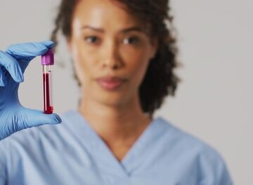 A healthcare worker wearing blue scrubs and protective blue gloves holding a glass collection tube filled with a red liquid sample and topped with a purple closure.