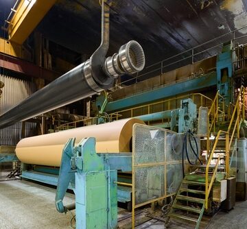 A large industrial paper machine in operation at a paper mill, featuring a massive finished paper roll and a heavy steel roll suspended by an overhead crane during maintenance or replacement.