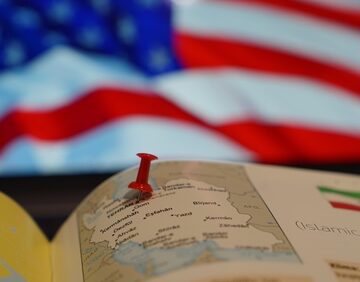 Image of a pushpin placed on a map of Iran with an American flag in the background.