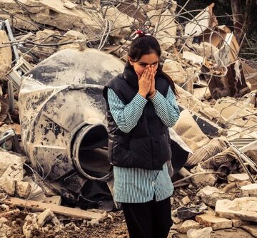 A person standing amidst heavy debris and wreckage, holding their hands to their face in a gesture of distress or grief. A person standing amidst heavy debris and wreckage, holding their hands to their face in a gesture of distress or grief.