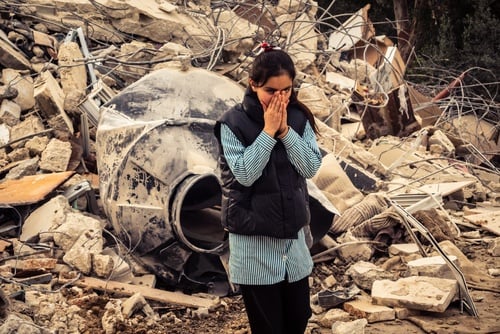 A person standing amidst heavy debris and wreckage, holding their hands to their face in a gesture of distress or grief. A person standing amidst heavy debris and wreckage, holding their hands to their face in a gesture of distress or grief.