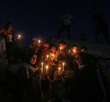 A group of Palestinian children and residents holding lit candles during a nighttime gathering or protest near damaged buildings in Rafah, in the southern Gaza Strip, on May 25, 2021. A group of Palestinian children and residents holding lit candles during a nighttime gathering or protest near damaged buildings in Rafah, in the southern Gaza Strip, on May 25, 2021.