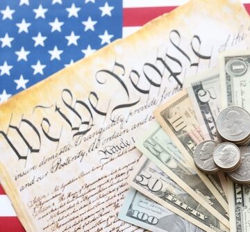 A close-up composition featuring U.S. currency (paper bills and stacked coins) resting on top of a replica of the U.S. Constitution (showing the "We the People" preamble) with an American flag background.