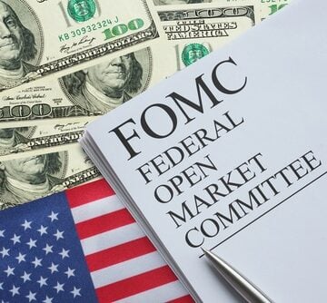 The image displays a document labeled FOMC (Federal Open Market Committee) surrounded by U.S. hundred-dollar bills and the American flag. The image displays a document labeled FOMC (Federal Open Market Committee) surrounded by U.S. hundred-dollar bills and the American flag.