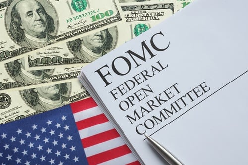 The image displays a document labeled FOMC (Federal Open Market Committee) surrounded by U.S. hundred-dollar bills and the American flag. The image displays a document labeled FOMC (Federal Open Market Committee) surrounded by U.S. hundred-dollar bills and the American flag.