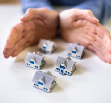 Concept of home protection, property security, or real estate asset management, featuring human hands shielding a group of miniature model houses.