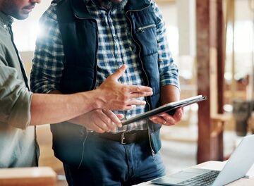 Two professionals review production plans and discuss manufacturing details in a workshop using a digital tablet and laptop. Two professionals review production plans and discuss manufacturing details in a workshop using a digital tablet and laptop.