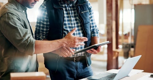Two professionals review production plans and discuss manufacturing details in a workshop using a digital tablet and laptop. Two professionals review production plans and discuss manufacturing details in a workshop using a digital tablet and laptop.