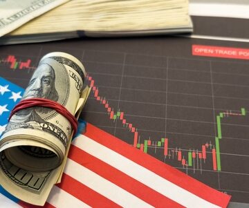 Concept image of a rolled-up bundle of U.S. hundred-dollar bills resting on an American flag, positioned against a background showing a declining stock market chart. Concept image of a rolled-up bundle of U.S. hundred-dollar bills resting on an American flag, positioned against a background showing a declining stock market chart.