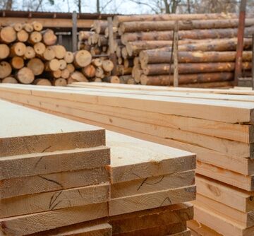 A commercial lumberyard with freshly cut, stacked dimensional softwood lumber in the foreground and stacked round logs in the background.