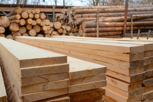 A commercial lumberyard with freshly cut, stacked dimensional softwood lumber in the foreground and stacked round logs in the background. A commercial lumberyard with freshly cut, stacked dimensional softwood lumber in the foreground and stacked round logs in the background.
