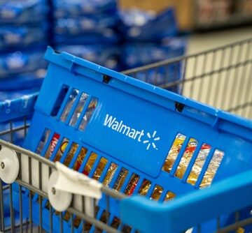 A blue Walmart shopping basket sits inside a wire shopping cart, holding items like pasta boxes on a store floor. A blue Walmart shopping basket sits inside a wire shopping cart, holding items like pasta boxes on a store floor.