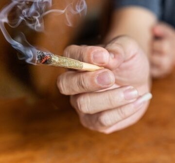 Finger,Holding,Lighted,Cannabis,Cigarette,With,Smoke,In,Dark,Room, AMERICANS ARE GETTING HIGH TO GET BY