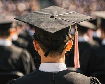 A rear view of a graduate wearing a black graduation cap (mortarboard) with a pink tassel and a dark academic gown during a commencement ceremony.