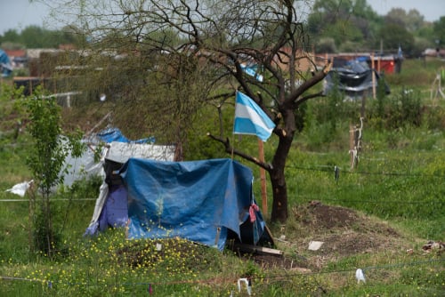 An informal housing or land occupation site in Guernica, Argentina, featuring an Argentine flag flying over a makeshift shelter.