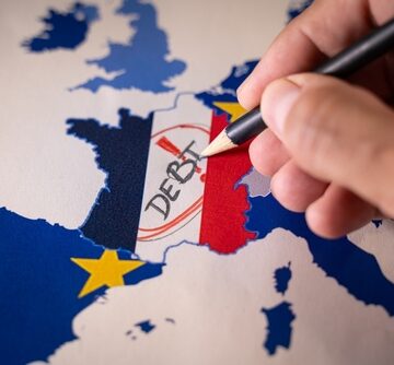 A hand with a pencil writing the word "DEBT" over a map of France colored with the French national flag, set against a map of Europe with EU star symbols. A hand with a pencil writing the word "DEBT" over a map of France colored with the French national flag, set against a map of Europe with EU star symbols.