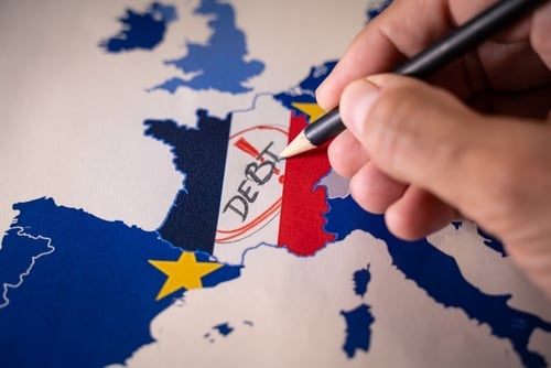 A hand with a pencil writing the word "DEBT" over a map of France colored with the French national flag, set against a map of Europe with EU star symbols. A hand with a pencil writing the word "DEBT" over a map of France colored with the French national flag, set against a map of Europe with EU star symbols.