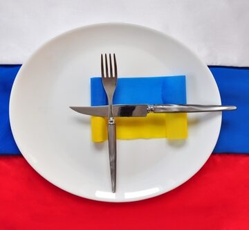 A plate and cutlery are set with a crossed silver fork and knife sitting on a background of the horizontal tricolor band of the Russian Federation flag and underneath the utensils is a smaller block representing the blue and yellow Ukrainian national flag. A plate and cutlery are set with a crossed silver fork and knife sitting on a background of the horizontal tricolor band of the Russian Federation flag and underneath the utensils is a smaller block representing the blue and yellow Ukrainian national flag.