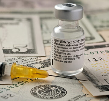 The image shows a Pfizer-BioNTech COVID-19 vaccine vial and a syringe resting on top of US dollar bills. The image shows a Pfizer-BioNTech COVID-19 vaccine vial and a syringe resting on top of US dollar bills.