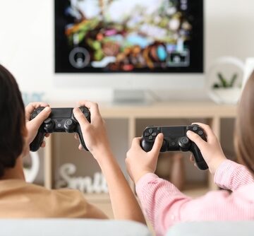 An image of two people sitting on a couch from behind, playing a video game on a TV using black wireless controllers that appear to resemble Sony DualShock 4 or DualSense style gamepads. An image of two people sitting on a couch from behind, playing a video game on a TV using black wireless controllers that appear to resemble Sony DualShock 4 or DualSense style gamepads.