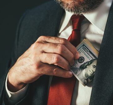 A man in a suit and red tie pulling a wad of U.S. $100 dollar bills from his jacket pocket.