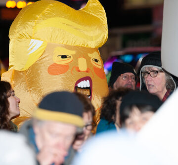A person wearing an inflatable, large-headed caricature with characteristic bright yellow hair and facial features resembling Donald Trump during a public demonstration.