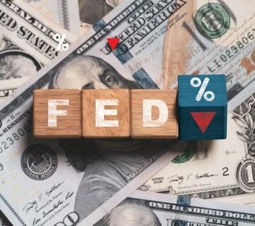 Illustration displays wooden letter blocks spelling out "FED" alongside a blue block marked with a percentage sign (%) and a prominent red downward-pointing arrow sitting directly on top of a scattered pile of United States one-hundred-dollar bills.