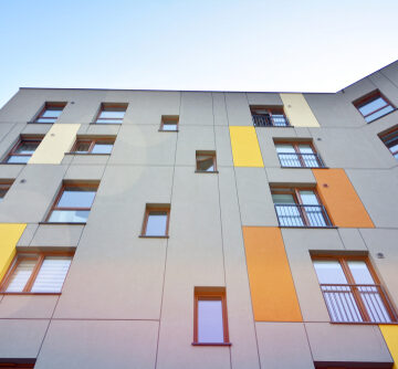 Image of the facade of a modern residential apartment building with grey paneling and colorful yellow and orange accents. Image of the facade of a modern residential apartment building with grey paneling and colorful yellow and orange accents.