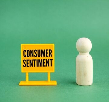 A white wooden figure stands next to a yellow sign reading "Consumer Sentiment." A white wooden figure stands next to a yellow sign reading "Consumer Sentiment."