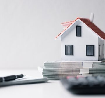The image shows a miniature white model house with a red roof resting on top of stacks of money, next to a calculator and pens on a white desk. The image shows a miniature white model house with a red roof resting on top of stacks of money, next to a calculator and pens on a white desk.