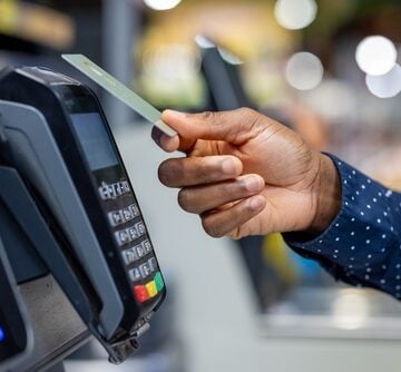 The image shows a person making a contactless payment by tapping a card on a point-of-sale (POS) payment terminal.