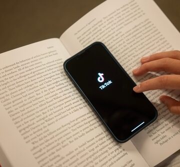 A smartphone displaying the TikTok app resting on top of an open book. A smartphone displaying the TikTok app resting on top of an open book.