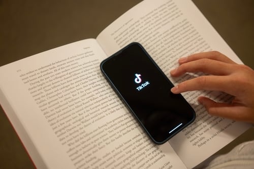 A smartphone displaying the TikTok app resting on top of an open book. A smartphone displaying the TikTok app resting on top of an open book.