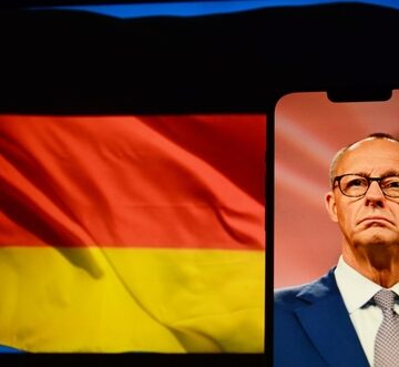 An image of German Chancellor Friedrich Merz displayed on a smartphone screen next to the German flag. An image of German Chancellor Friedrich Merz displayed on a smartphone screen next to the German flag.