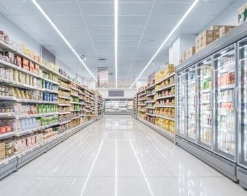A long, empty, brightly lit modern supermarket aisle lined with stocked grocery shelves on the left and glass-door refrigerator and freezer cases on the right. A long, empty, brightly lit modern supermarket aisle lined with stocked grocery shelves on the left and glass-door refrigerator and freezer cases on the right.