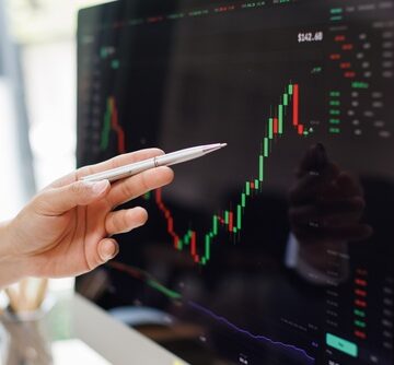 A hand holding a pen is pointing at a financial chart or stock market candlestick graph on a computer monitor. A hand holding a pen is pointing at a financial chart or stock market candlestick graph on a computer monitor.