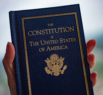 A person holding a small blue pocket-sized hardcover edition of The Constitution of the United States of America. A person holding a small blue pocket-sized hardcover edition of The Constitution of the United States of America.