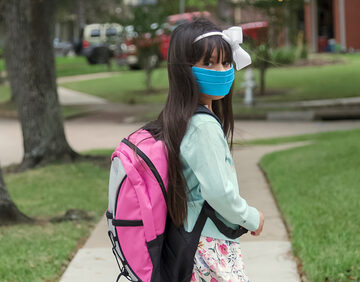 A young student wearing a blue face mask and a pink backpack while walking outdoors on a sidewalk near trees and a building. A young student wearing a blue face mask and a pink backpack while walking outdoors on a sidewalk near trees and a building.