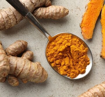 The image shows turmeric (Curcuma longa), featuring whole raw roots (rhizomes), bright orange sliced pieces, and a spoon filled with ground golden turmeric powder. The image shows turmeric (Curcuma longa), featuring whole raw roots (rhizomes), bright orange sliced pieces, and a spoon filled with ground golden turmeric powder.