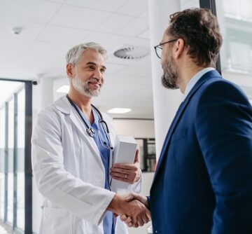 A male doctor in a white coat with a stethoscope shaking hands with a businessman in a suit in a hospital or clinic hallway.