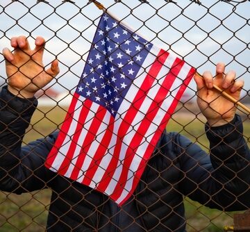 Photo of a person wearing a dark jacket gripping a chain-link fence with both hands while holding a small U.S. flag. Photo of a person wearing a dark jacket gripping a chain-link fence with both hands while holding a small U.S. flag.