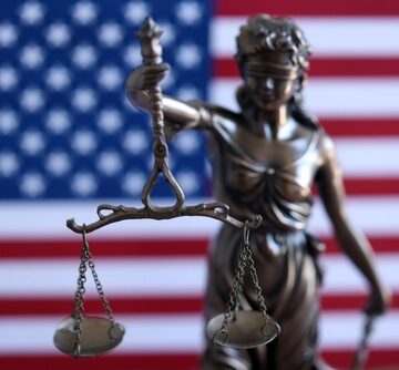 A bronze-toned statue of Lady Justice holding the scales of justice in front of an American flag.