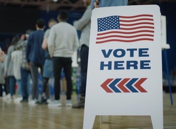 A line of people stand behind a "Vote Here" sign at an in-person polling place. A line of people stand behind a "Vote Here" sign at an in-person polling place.
