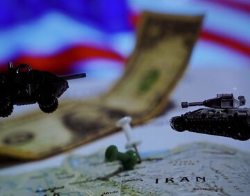Miniature military vehicles placed over a map of Iran, with a U.S. dollar curved across the display, and a blurred American flag visible in the background.